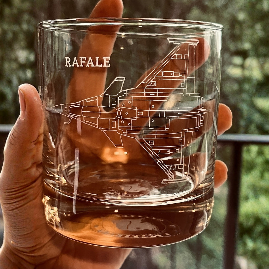 Clear glass mug with 'RAFALE' aircraft engraving held by a hand against a blurred green background