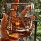 Clear glass mug with 'RAFALE' aircraft engraving held by a hand against a blurred green background