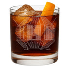 A clear rocks glass filled with an amber-brown cocktail, ice, and an orange peel garnish, shown on a white background. The glass features an etched white line drawing of india's irst satellite - the aryabhata from ISRO