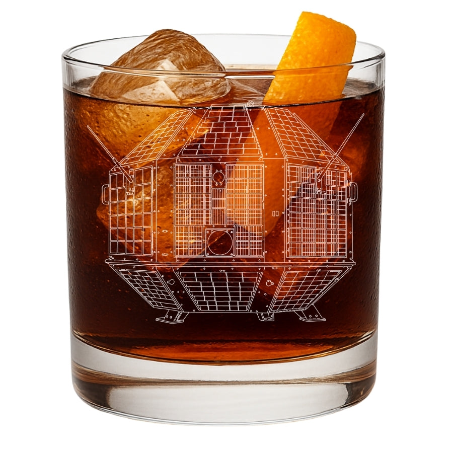 A clear rocks glass filled with an amber-brown cocktail, ice, and an orange peel garnish, shown on a white background. The glass features an etched white line drawing of india's irst satellite - the aryabhata from ISRO