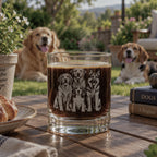 A clear glass tumbler with an engraved portrait of four dogs sits on a wooden outdoor table. The background shows a sunny backyard with two dogs lying on the grass, a potted plant, and books on the table.