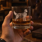A hand holds a clear glass rocks tumbler engraved with a Golden Retriever dog and the words 'Golden Retriever' on the front, filled with amber whiskey in a dimly lit, warm indoor setting.