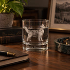 A clear glass rocks tumbler with an etched Golden Retriever dog and the words 'Golden Retriever' on the base, displayed on a wooden desk with books, a plant, and framed picture in the background.