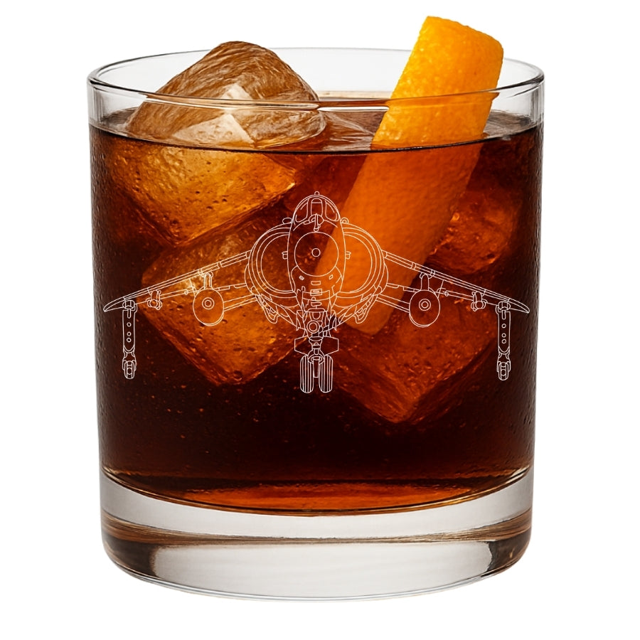 A clear rocks glass filled with an amber-brown cocktail, large ice cubes, and an orange peel garnish, shown on a white background. The glass is etched with a detailed white line drawing of a hawker harrier fighter jet.