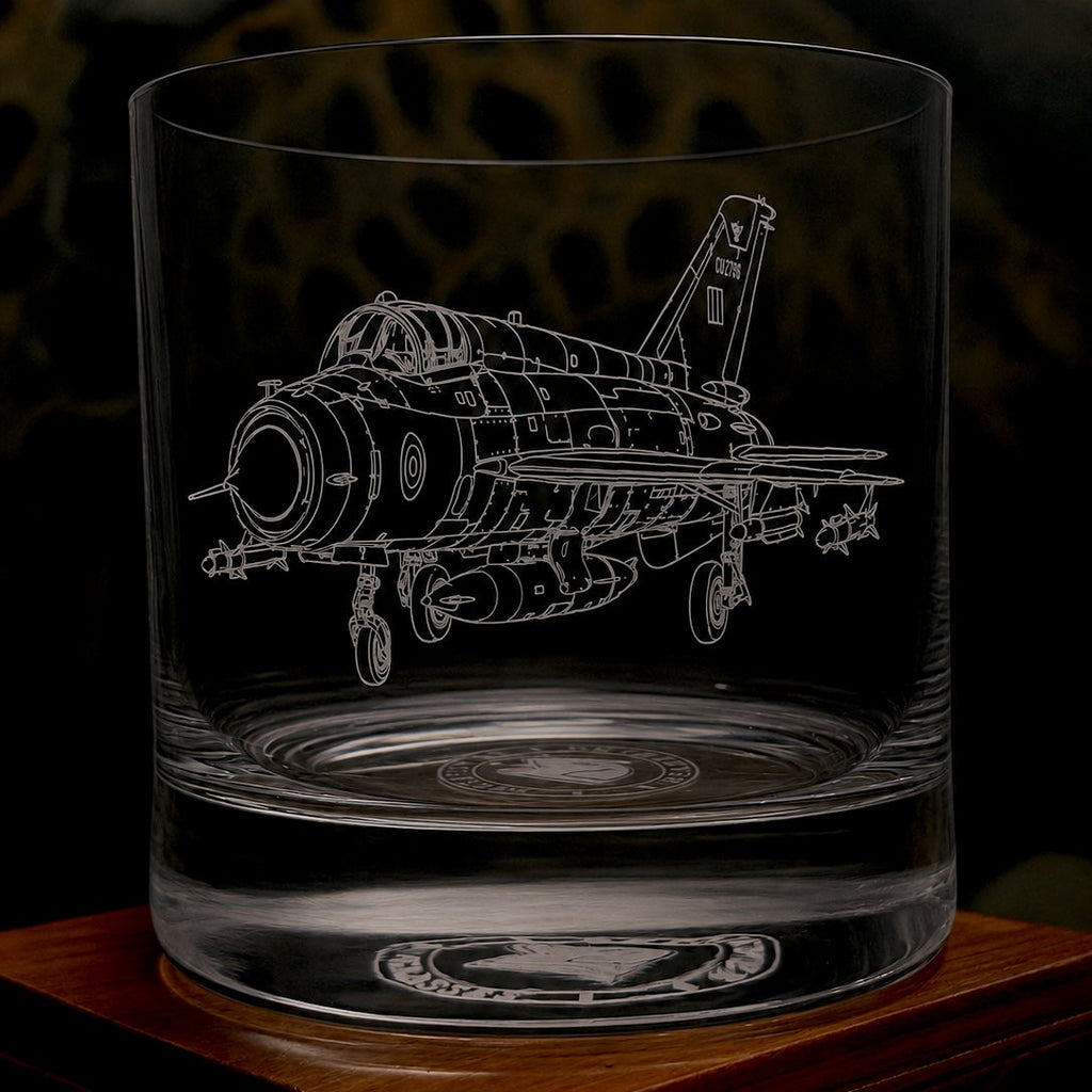 A clear glass tumbler with a detailed white etched line drawing of a Mig-21 military fighter jet, shown on a dark background with a wooden surface.