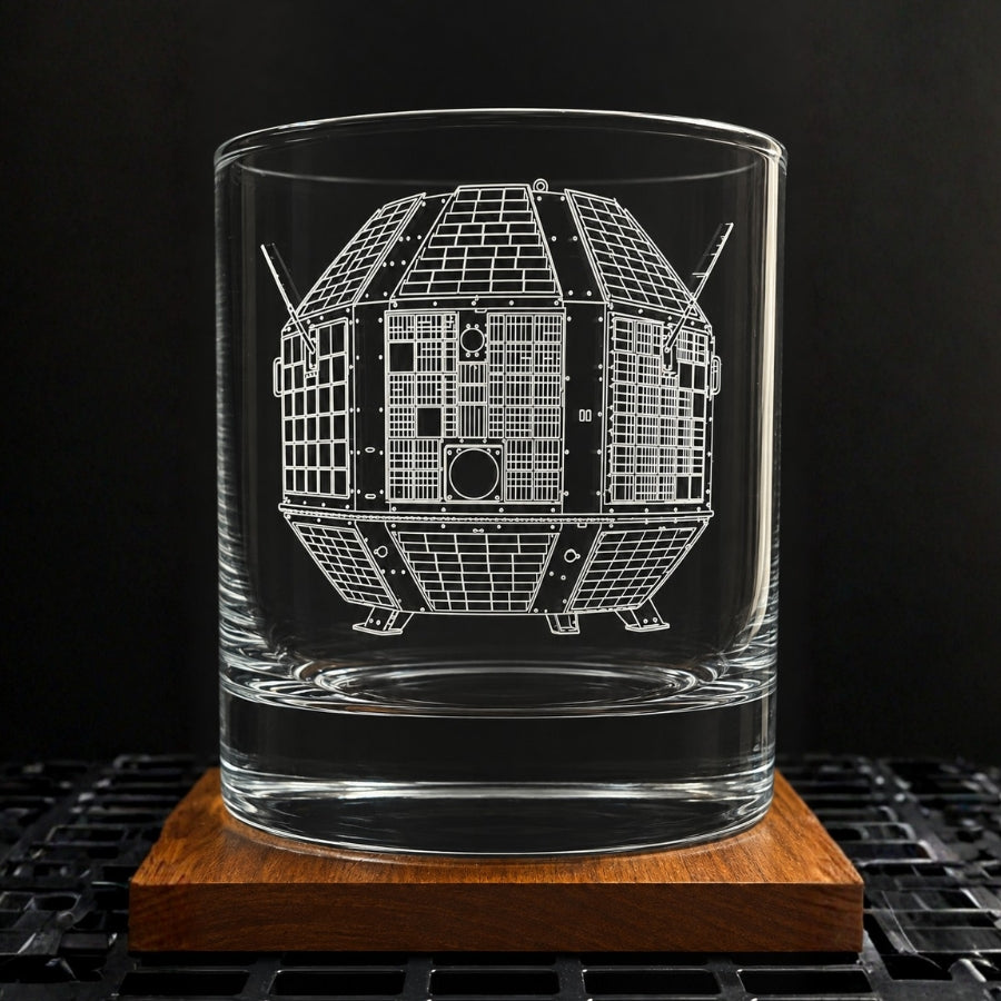 A clear glass rocks tumbler with an etched white schematic of the indian aryabhata satellite, displayed on a square wooden coaster against a dark background.